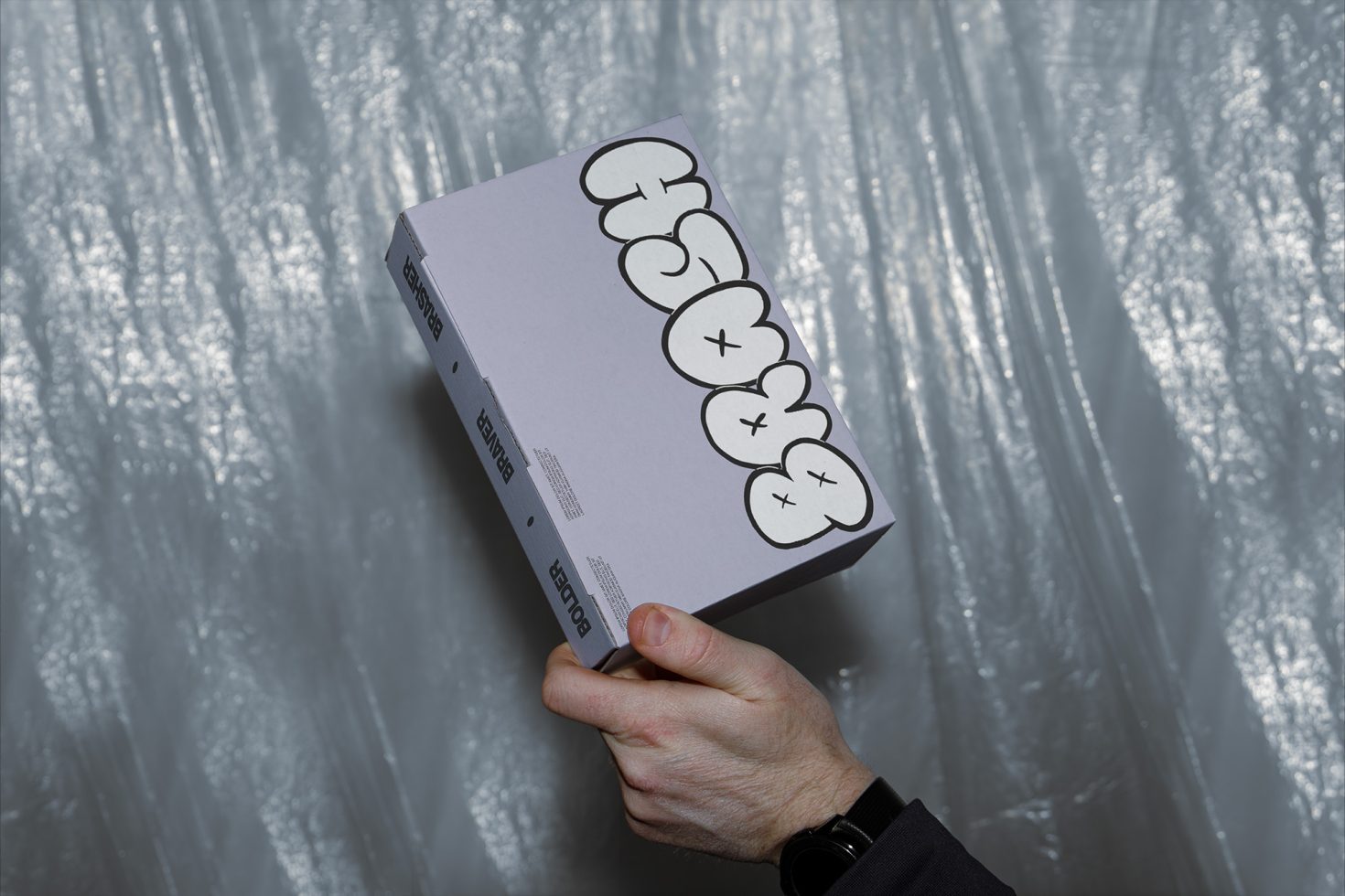 Person holding a box with graphic design mockup, silver texture background, packaging design example, product presentation.